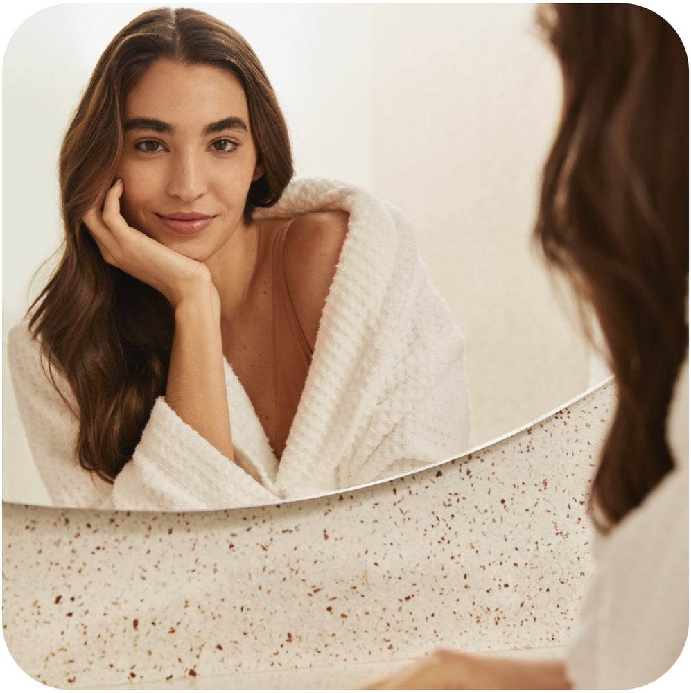 What Causes Oily Hair: 9 Reasons & Solutions Woman in a white bathrobe looking at her reflection in a mirror while wondering what causes oily hair