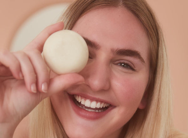 Smiling woman with blonde hair holding one of Bars Over Bottles shampoo bars over her right eye