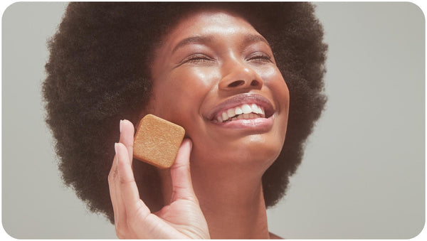 A smiling curly haired woman holding a Bars Over Bottles Facial Cleansing Bar for Balanced Skin close to her face