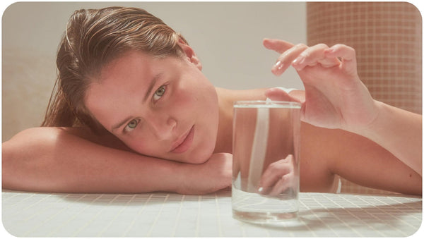 A glass of water in front of a woman with dark blonde hair who uses Bars Over Bottles no poo shampoo