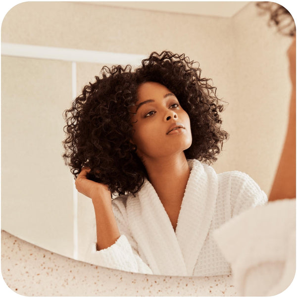 Woman in a white robe is looking in a mirror while touching her soft coily hair after using shampoo and conditioner bars for curly hair