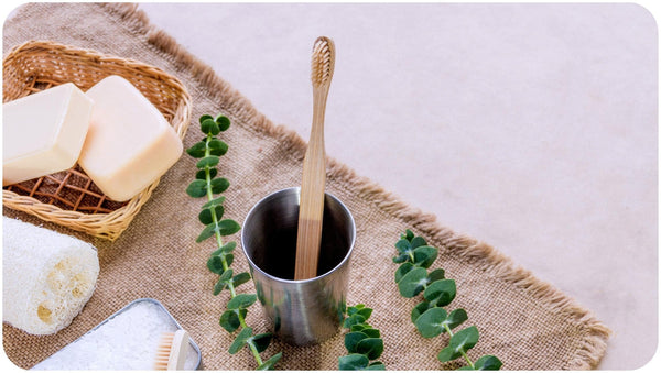 Bar soaps in a basket and a wooden brush in a stainless cup placed on a vintage linen tablecloth signifying waterless beauty